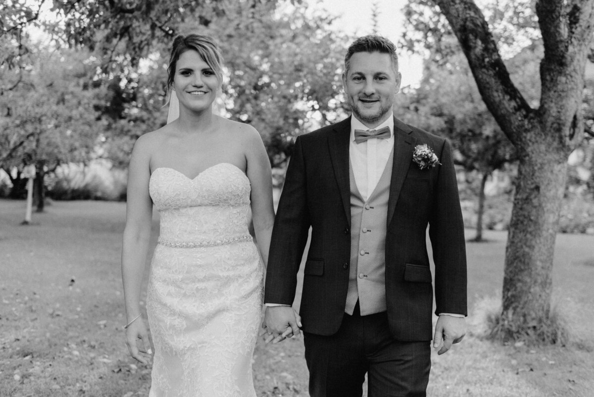 christian-lorenz.net Braut und Bräutigam stehen Händchen haltend in einem Park, umgeben von Bäumen. Die Braut trägt ein trägerloses Kleid, der Bräutigam einen Anzug mit Fliege.