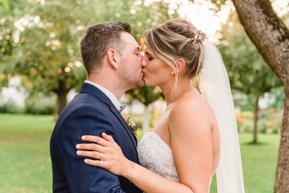 christian-lorenz.net Braut und Bräutigam küssen sich im Freien in einem Garten. Der Schleier der Braut weht, der Bräutigam trägt einen dunklen Anzug. Im Hintergrund sind Bäume und grünes Gras zu sehen.