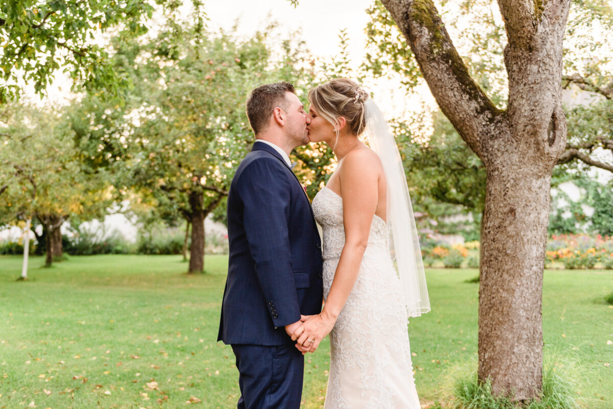 christian-lorenz.net Braut und Bräutigam küssen sich Händchen haltend in einem Garten. Die Braut trägt ein weißes Kleid und einen Schleier, der Bräutigam einen marineblauen Anzug. Bäume und Gras umgeben sie.