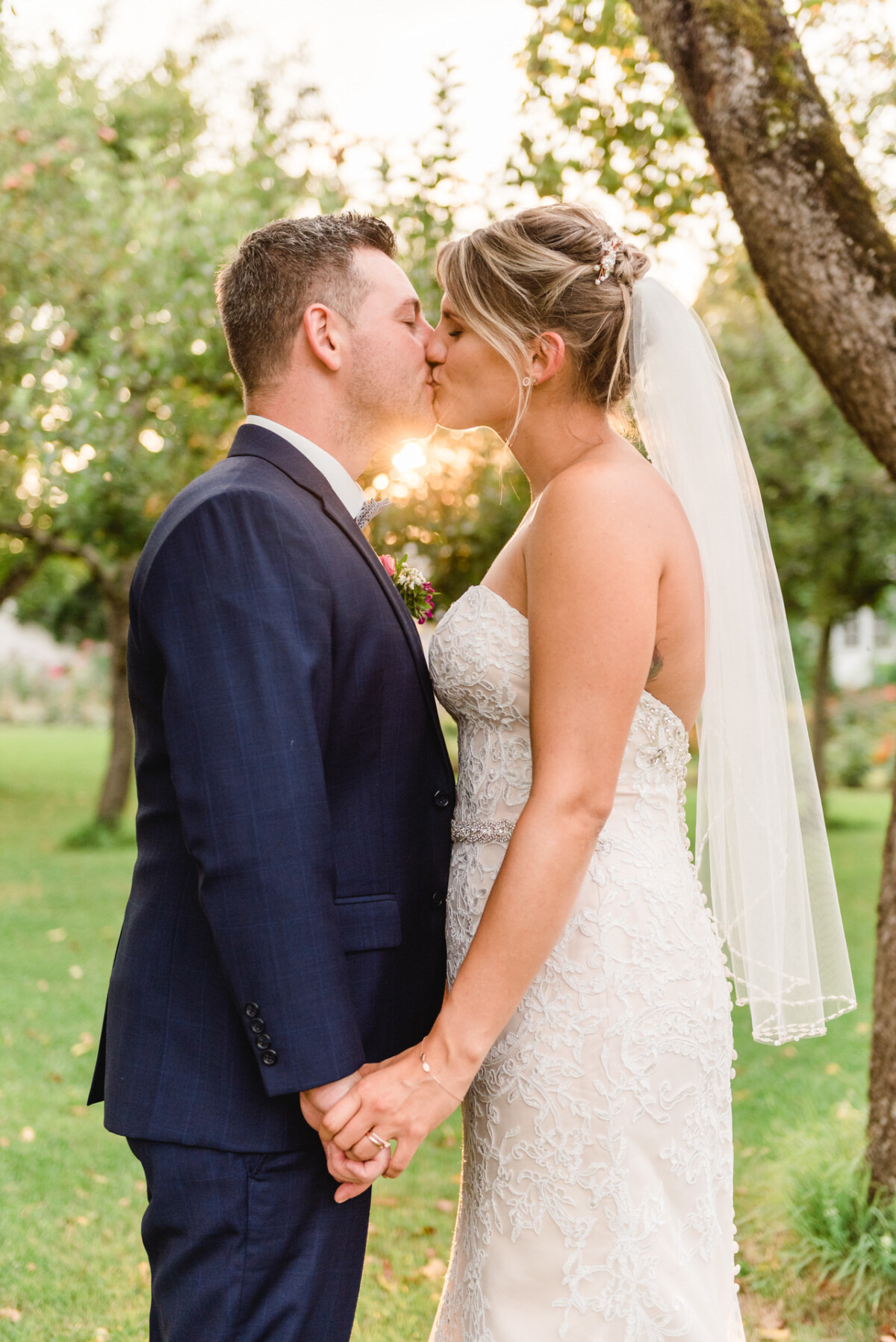 christian-lorenz.net Braut und Bräutigam küssen sich im Freien unter einem Baum und halten sich an den Händen. Sonnenlicht fällt durch die Zweige und beleuchtet sie.