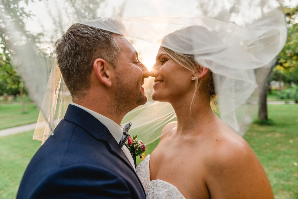 christian-lorenz.net Braut und Bräutigam lächeln sich unter einem wallenden Schleier im Freien an, während das Sonnenlicht durchscheint.