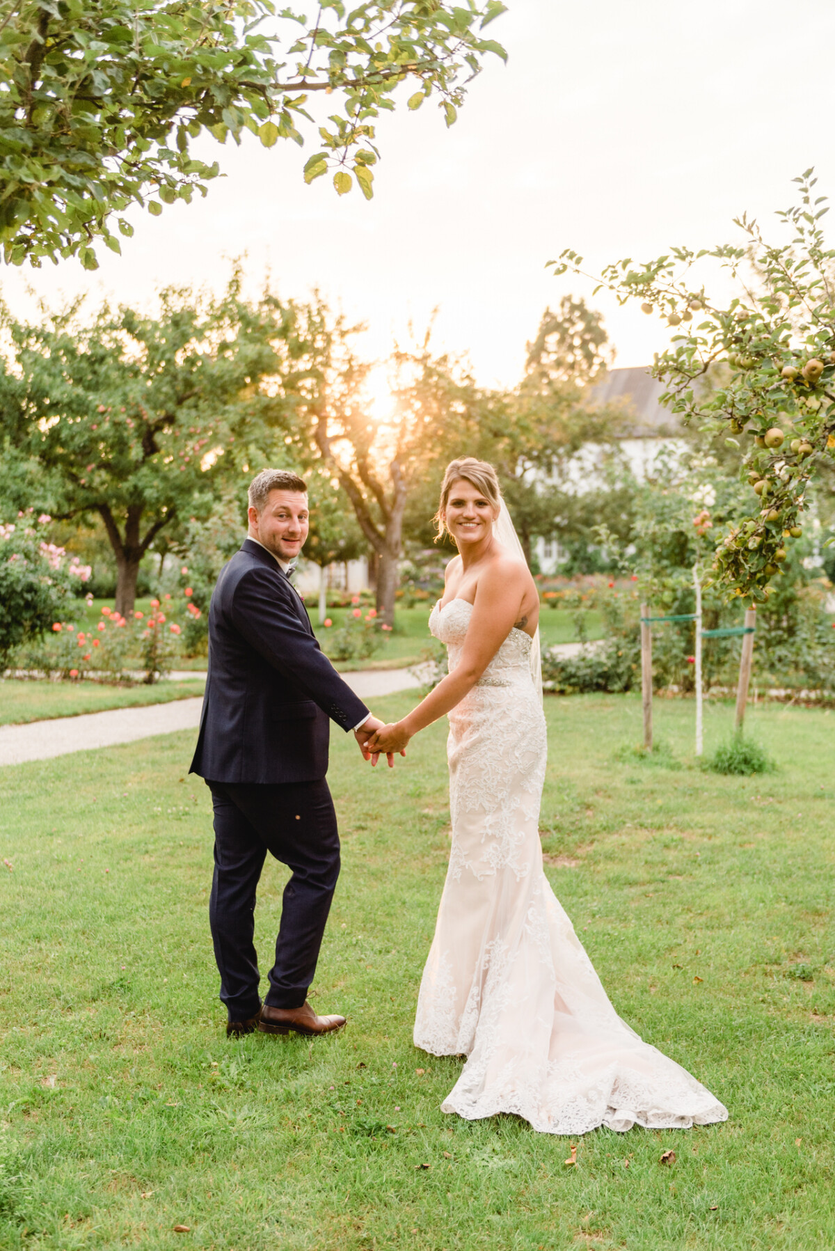 christian-lorenz.net Eine Braut und ein Bräutigam stehen Hand in Hand auf einer Wiese mit Bäumen und blicken zurück, während im Hintergrund die Sonne untergeht.