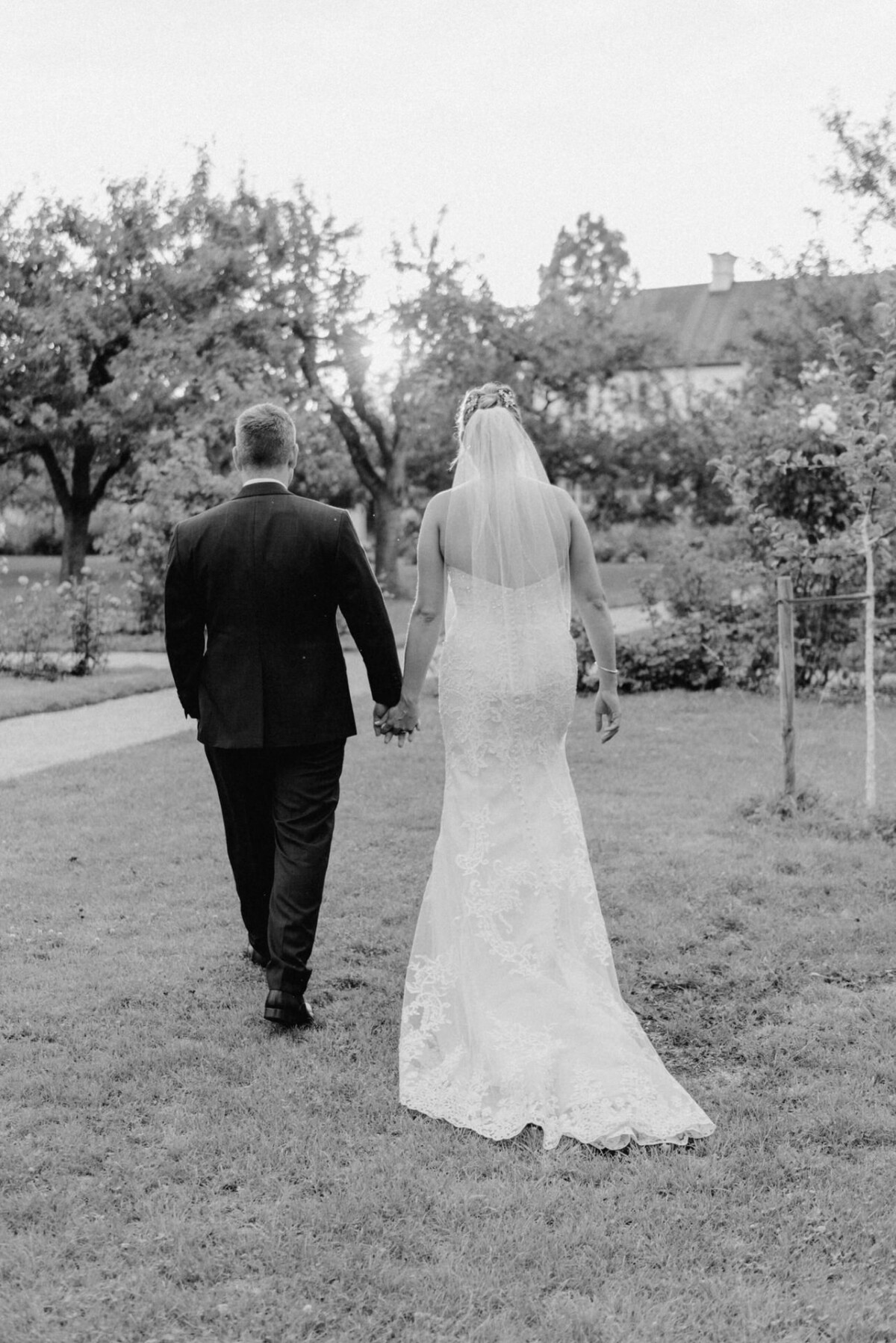 christian-lorenz.net Braut und Bräutigam gehen Hand in Hand auf dem Gras, mit dem Gesicht abgewandt, mit Bäumen und einem Haus im Hintergrund.