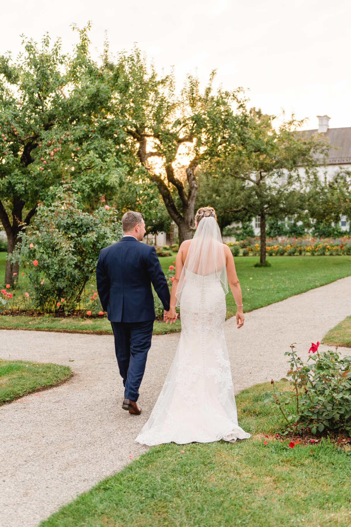 christian-lorenz.net Eine Braut und ein Bräutigam gehen Hand in Hand einen Gartenweg entlang, umgeben von Bäumen und Blumen, mit einem großen Gebäude im Hintergrund.
