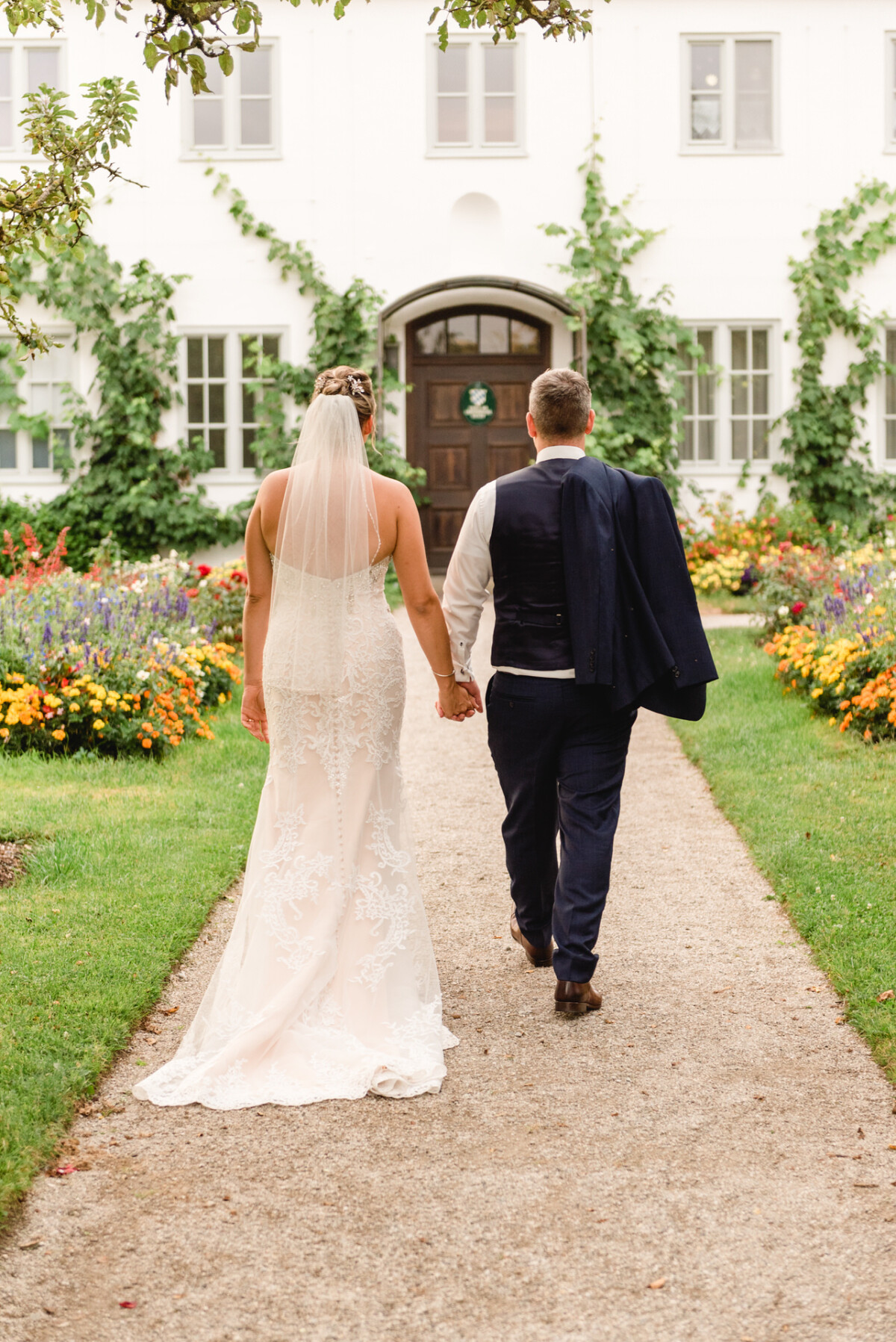 christian-lorenz.net Ein Paar in Hochzeitskleidung geht Hand in Hand auf ein Gebäude mit einem Gartenweg zu, der von bunten Blumen und Grün umgeben ist.