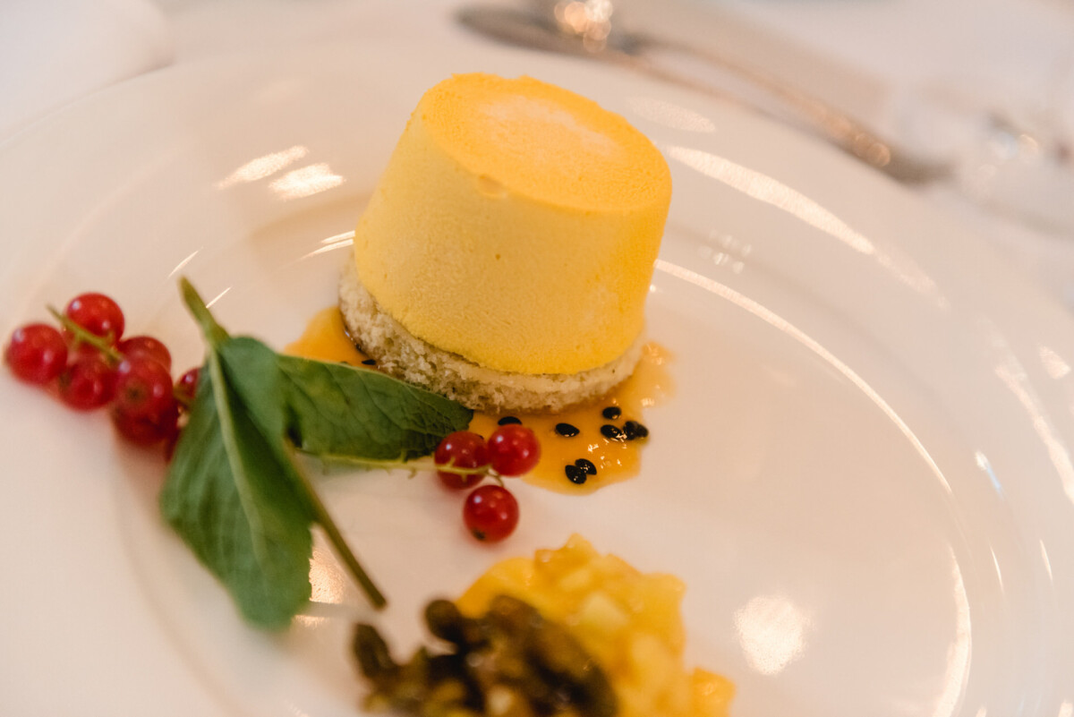 christian-lorenz.net Ein rundes Zitronenmousse-Dessert auf einem Teller, garniert mit roten Johannisbeeren, Blättern und einer kleinen Portion Soße.