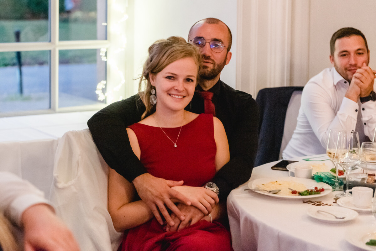 christian-lorenz.net Eine Frau in einem roten Kleid sitzt auf dem Schoß eines Mannes, beide lächeln, an einem Esstisch mit Tellern und Weingläsern; ein weiterer Mann in einem weißen Hemd sitzt in der Nähe.