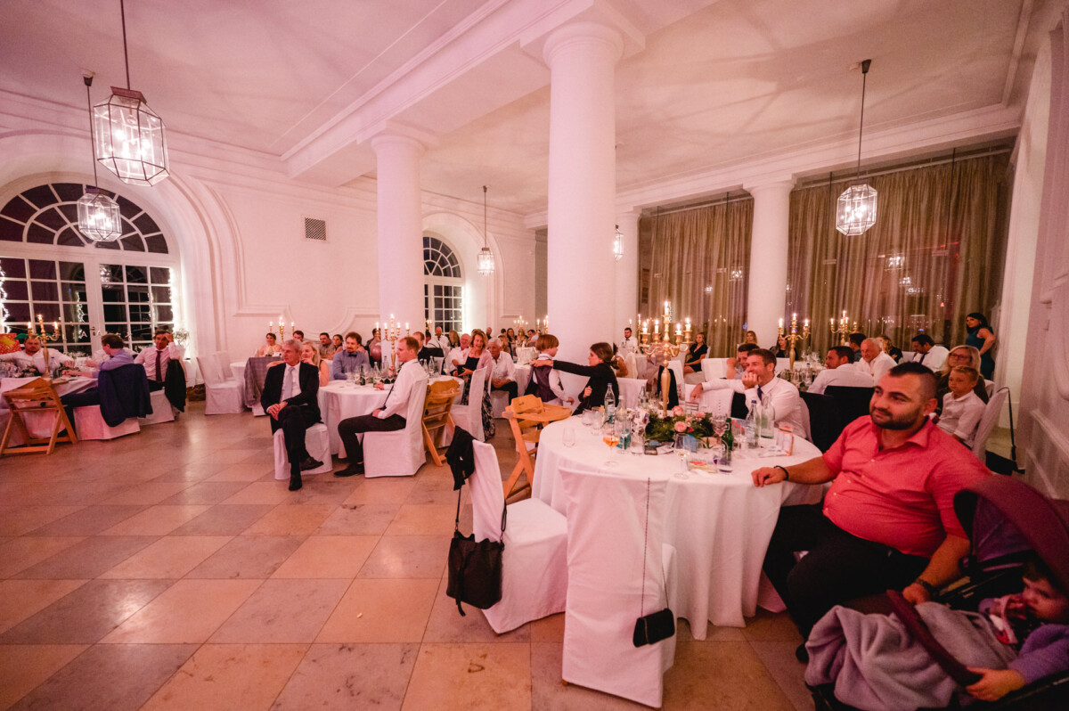 christian-lorenz.net Menschen sitzen an runden Tischen in einem dekorierten Bankettsaal mit weißen Säulen und großen Fenstern und hören aufmerksam zu.