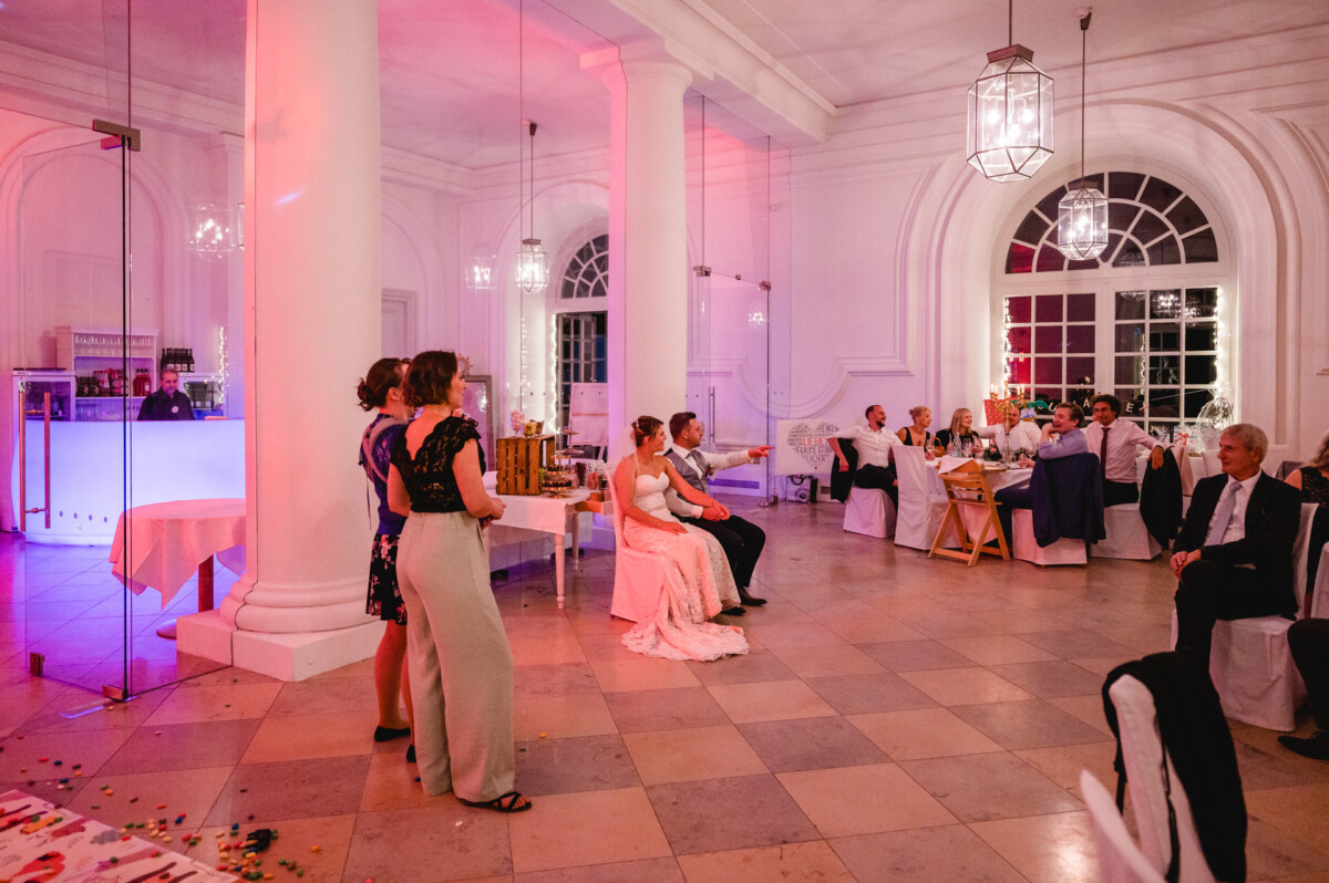 christian-lorenz.net Hochzeitsempfang in einem festlichen Saal mit sitzenden und stehenden Gästen. Braut und Bräutigam stehen im Mittelpunkt. Der Raum ist mit rosa und weißen Lichtern beleuchtet und verfügt über große Fenster und Säulen.