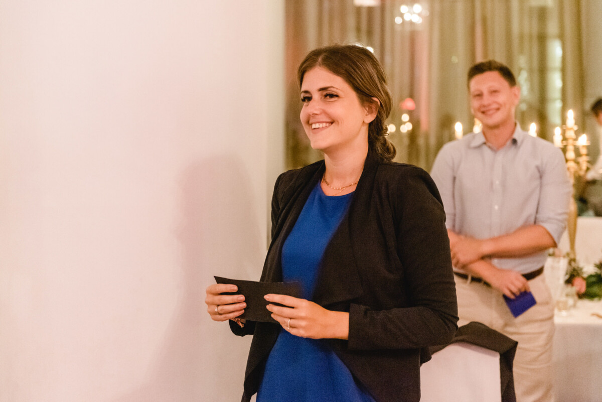 christian-lorenz.net Eine Frau in blauem Kleid und schwarzem Blazer lächelt und hält einen Umschlag in der Hand. Im Hintergrund lächelt ein Mann in hellem Hemd. Sie befinden sich in einem dekorierten Raum mit Tischen und Kerzen.