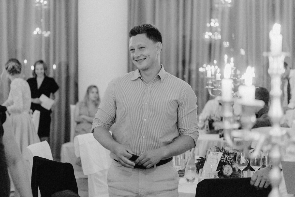 christian-lorenz.net Eine lächelnde Person steht bei einer formellen Veranstaltung und hält ein Gerät in der Hand. Im Hintergrund sind Tische, Menschen und Kandelaber zu sehen. Die Kulisse ist elegant und dekorativ.