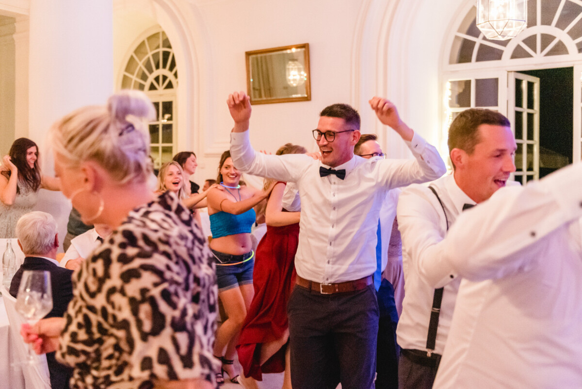 christian-lorenz.net In einem hell erleuchteten Raum mit großen Fenstern wird getanzt und gefeiert. Ein Mann mit Fliege reckt die Arme in die Höhe, während andere um ihn herum tanzen.