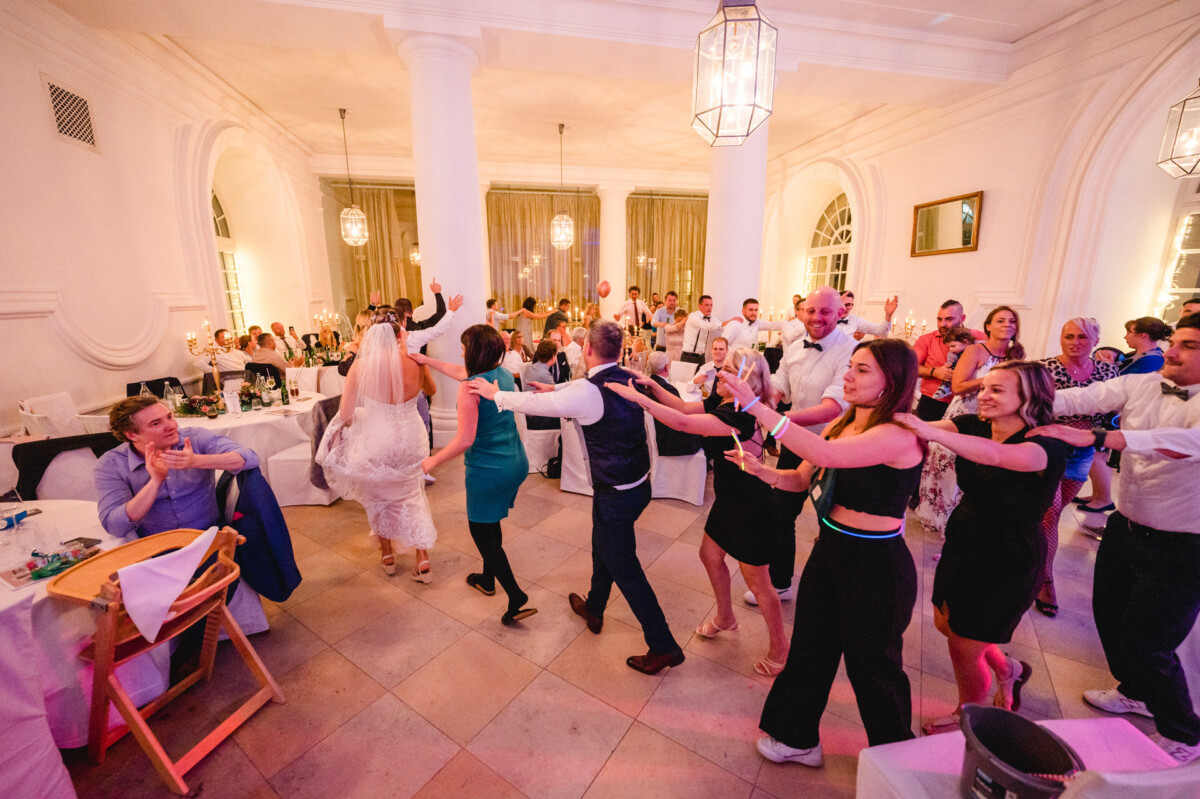christian-lorenz.net Bei einem Hochzeitsempfang tanzen die Gäste in einer Polonaise in einem üppig dekorierten Saal mit weißen Säulen und Kronleuchtern.