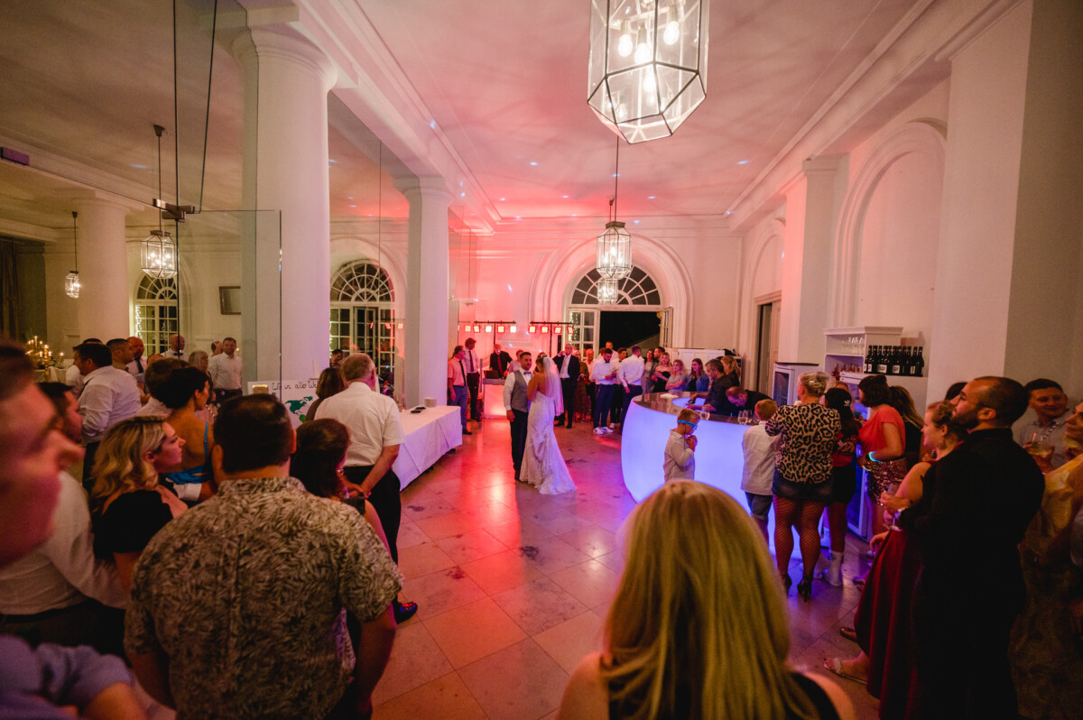 christian-lorenz.net Die Menschen versammelten sich in einem eleganten, hellen Raum mit Bogenfenstern. In der Mitte stand ein Paar, im Hintergrund ein DJ-Pult und Partylichter.