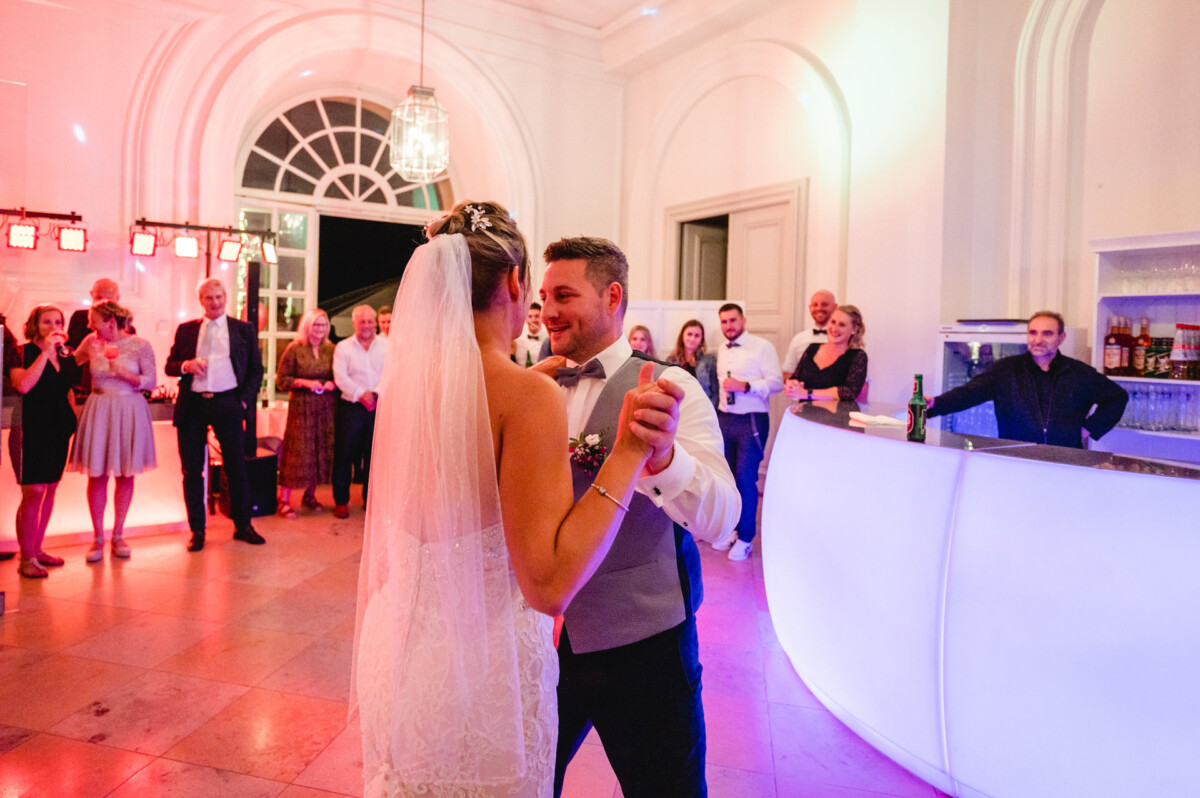 christian-lorenz.net Ein Brautpaar tanzt in einem hell erleuchteten Raum, umgeben von lächelnden Gästen. Im Hintergrund steht eine weiße Bar mit Getränken.