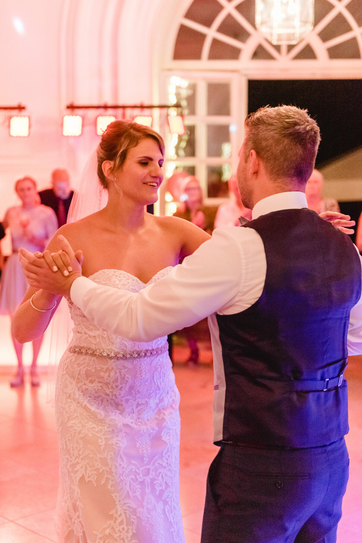 christian-lorenz.net Braut und Bräutigam tanzen in einem warm beleuchteten Ballsaal, umgeben von Gästen. Die Braut trägt ein weißes Kleid und der Bräutigam eine Weste.