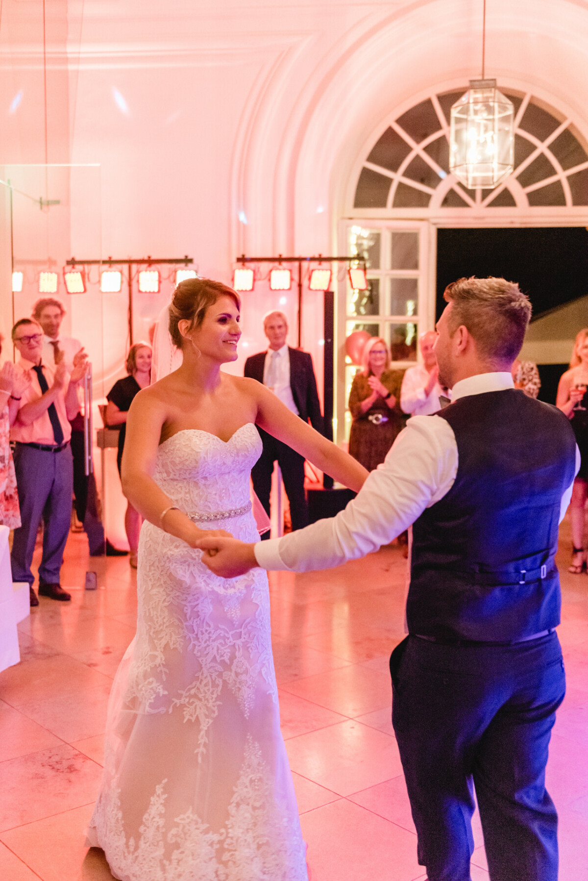 christian-lorenz.net Braut und Bräutigam tanzen zusammen in einem warm beleuchteten Veranstaltungsort, umgeben von Gästen.
