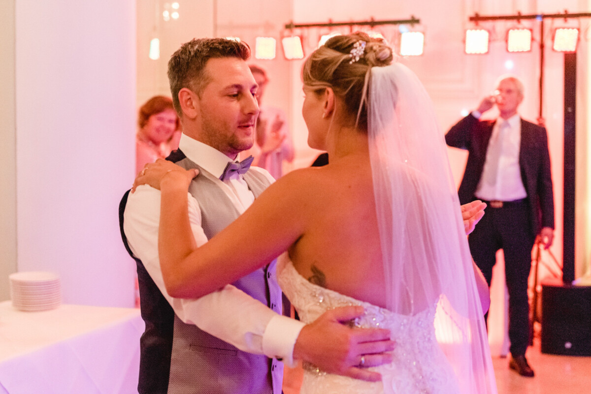 christian-lorenz.net Braut und Bräutigam tanzen gemeinsam den ersten Tanz auf ihrer Hochzeitsfeier. Sie trägt ein weißes Kleid mit Schleier, er eine Weste und Fliege. Im Hintergrund sind Gäste und ein Fotograf zu sehen.