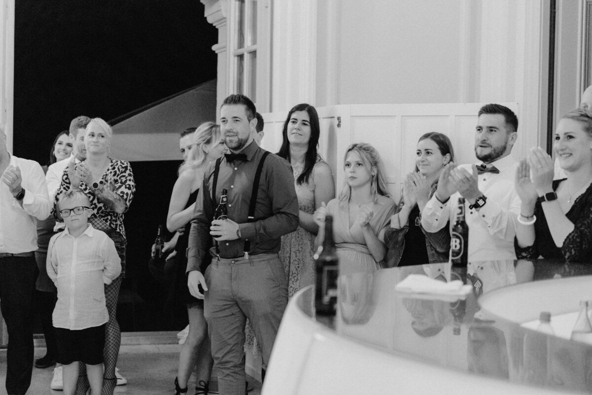 christian-lorenz.net Eine Gruppe von Menschen, darunter Männer, Frauen und ein Kind, steht in einem Veranstaltungsort und klatscht. Einige halten Getränke in der Hand, und alle sind formelle oder halbformelle Kleidung.