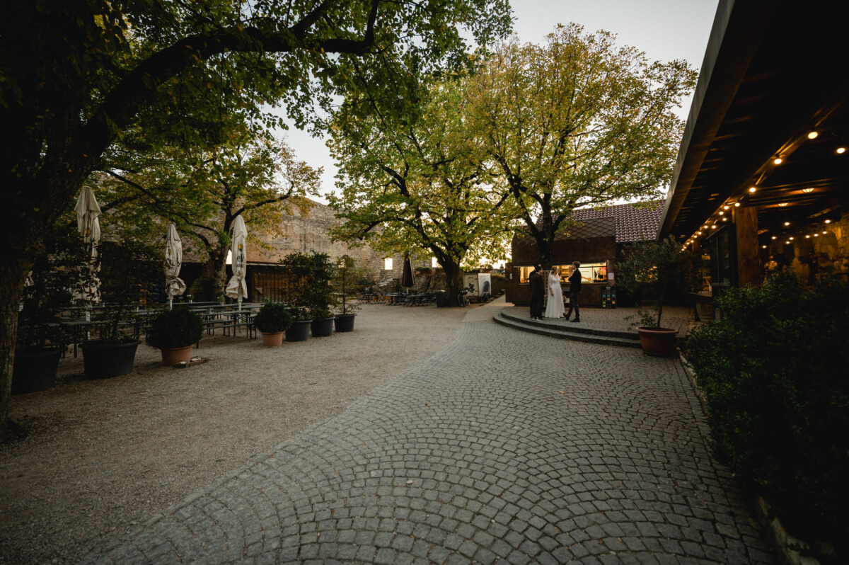 Ein Innenhof mit Kopfsteinpflasterwegen und hohen Bäumen. Links stehen Sonnenschirme und Tische, rechts ein Gebäude mit Lichterketten und Schrägdach. In der Ferne stehen Menschen.