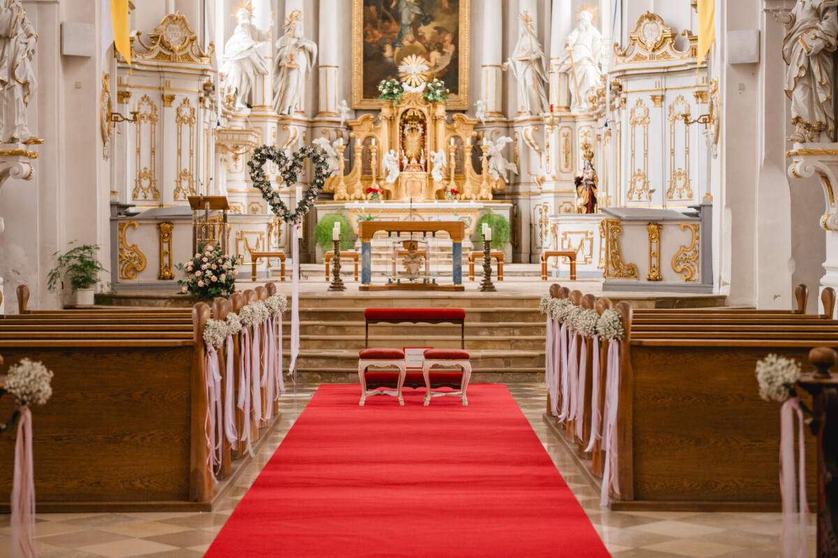 2024.10.05-Lea-Viktor-Hochzeit-Schammenhof-Langenau-bei-Ulm-00005 Der kunstvolle Kircheninnenraum in der Nähe der Hochzeit Schammenhof in Langenau bei Ulm verfügt über einen Gang mit rotem Teppich, Holzbänke, Blumendekor und zwei Kniebänke vor einem kunstvollen Altar, der mit Statuen und goldenen Akzenten geschmückt ist.