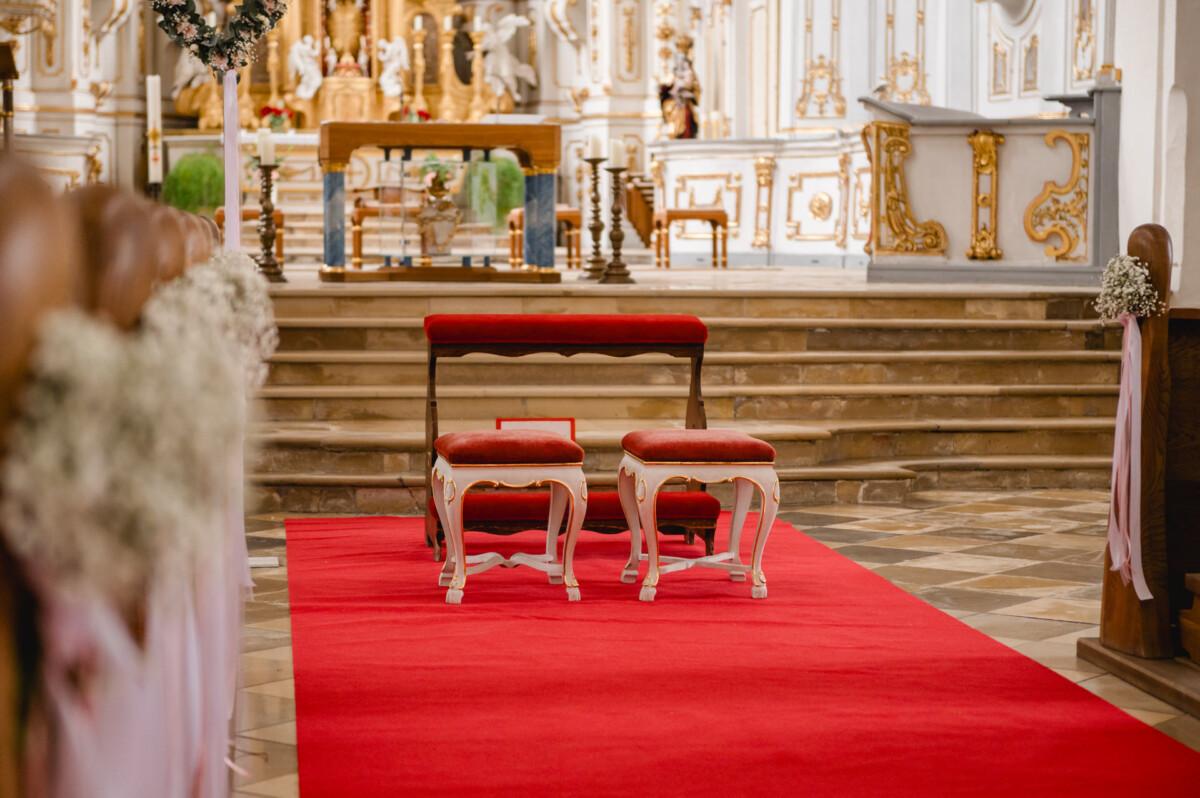 2024.10.05-Lea-Viktor-Hochzeit-Schammenhof-Langenau-bei-Ulm-00007 Zwei Kniebänke mit roten Kissen ruhen auf einem weichen Teppich in einem kunstvollen Kirchenraum, der mit goldenen Akzenten und Blumendekorationen geschmückt ist und den perfekten Rahmen für eine Hochzeit im charmanten Schammenhof Langenau bei Ulm bietet.