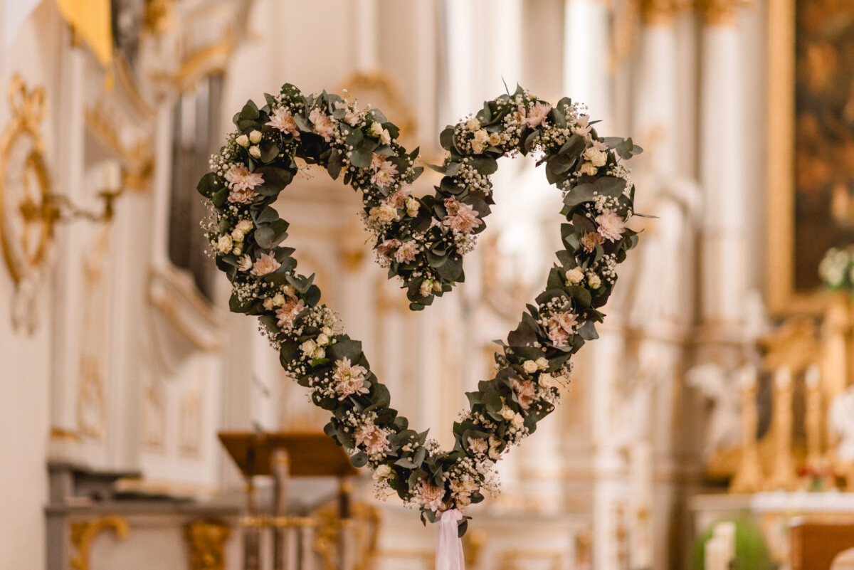 christian-lorenz.net Herzförmiger Blumenkranz mit Grünpflanzen und kleinen weißen Blüten vor dem kunstvollen Innenraum der Schammenhofkirche in Langenau bei Ulm.