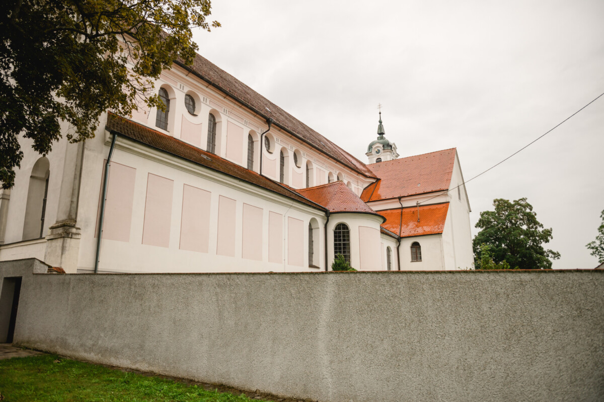 Eine weiß-rosa Kirche mit rotem Dach und Turm steht hinter einer grauen Mauer und grünem Rasen unter einem bewölkten Himmel.