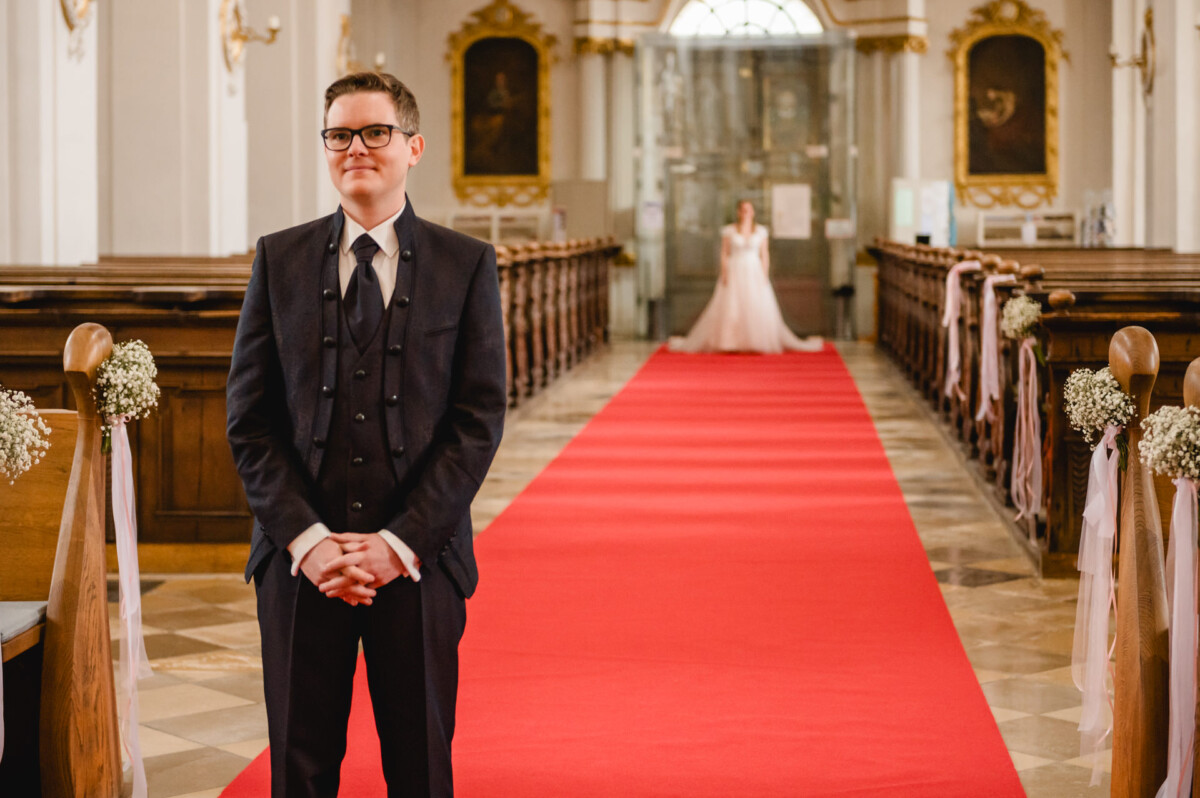 Ein Bräutigam steht in einer Kirche am Altar und wartet auf die Braut, die im Hintergrund durch einen mit rotem Teppich ausgelegten Gang schreitet.