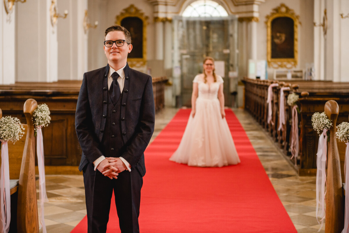 Eine Braut geht den Gang entlang auf einen Bräutigam zu, der lächelnd in einer Kirche mit rotem Teppich und Blumenschmuck steht.