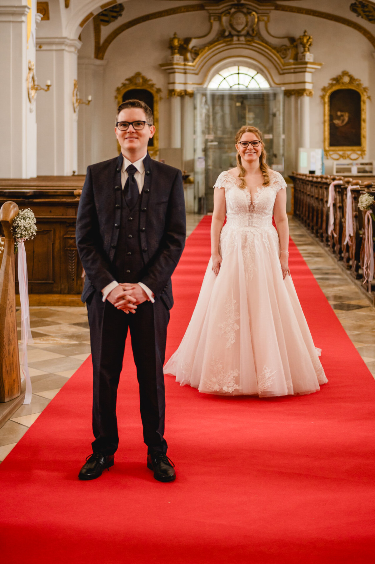 Braut und Bräutigam stehen auf einem roten Teppich in einer Kirche. Der Bräutigam im Anzug steht im Vordergrund, während die Braut im weißen Kleid lächelnd hinter ihm steht.