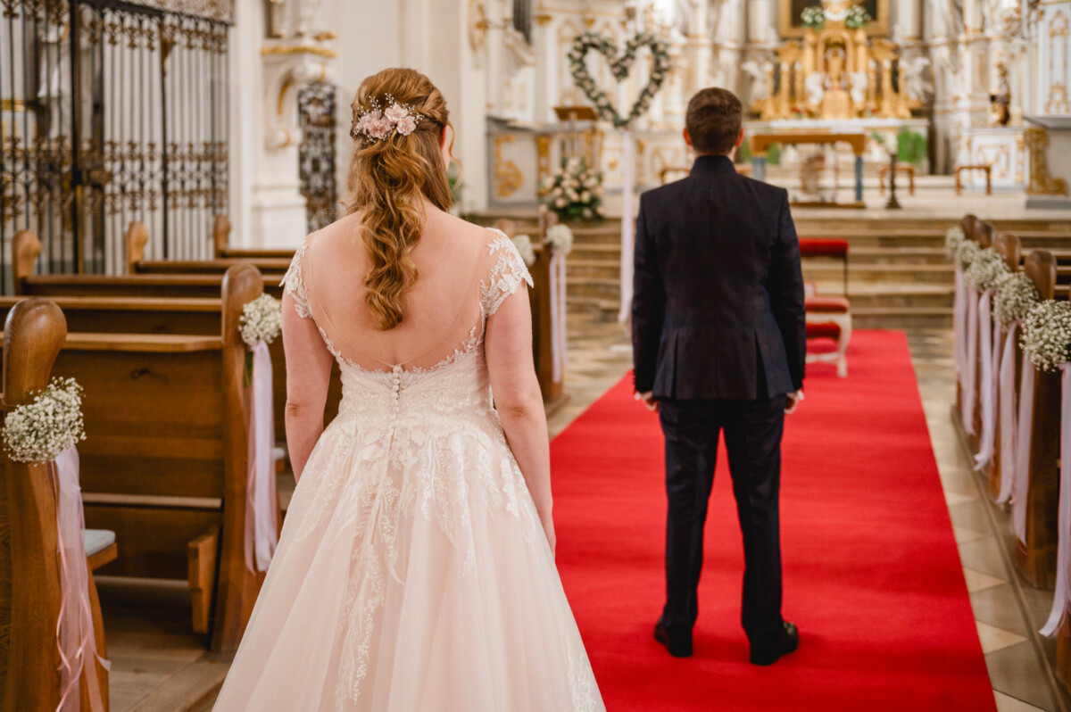 Eine Braut im weißen Kleid steht auf einem roten Teppich im Kirchengang. Ihr Bräutigam im dunklen Anzug steht ihr mit dem Rücken zu. Blumenschmuck schmückt die Kirchenbänke und den Altar.