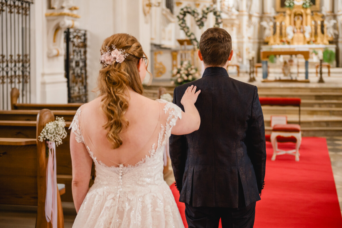 In einer Kirche mit kunstvollen Dekorationen und einem Gang mit rotem Teppich tippt eine Braut ihrem Bräutigam sanft auf die Schulter.