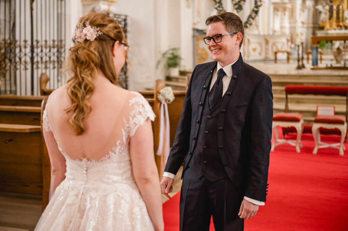 Eine Braut und ein Bräutigam lächeln sich in einer geschmückten Kirche an. Sie tragen ein weißes Brautkleid und einen dunklen Anzug.