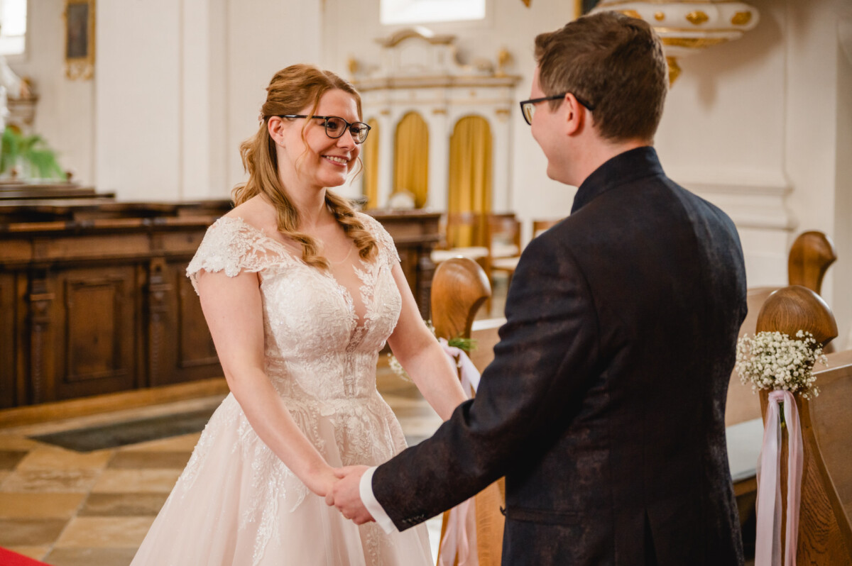 Eine Braut und ein Bräutigam halten sich an den Händen und lächeln sich in einer Kirche an, während sie neben einer Holzbank stehen.