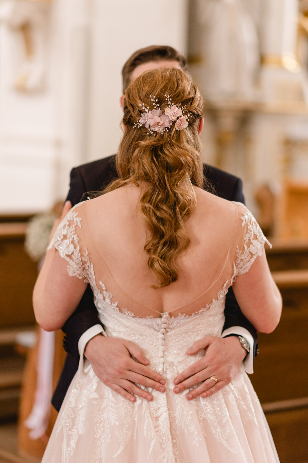 christian-lorenz.net Braut und Bräutigam umarmen sich und blicken einander zu. Die Braut steht mit dem Rücken zur Kamera und trägt ein Spitzenkleid und einen floralen Haarschmuck. Der Bräutigam hält ihre Taille in einer kirchlichen Umgebung.