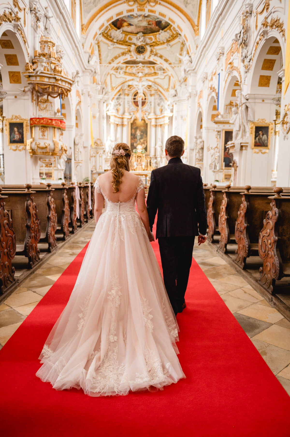 Ein Paar geht in Hochzeitskleidung in einer reich verzierten Kirche mit rotem Teppich den Gang entlang.