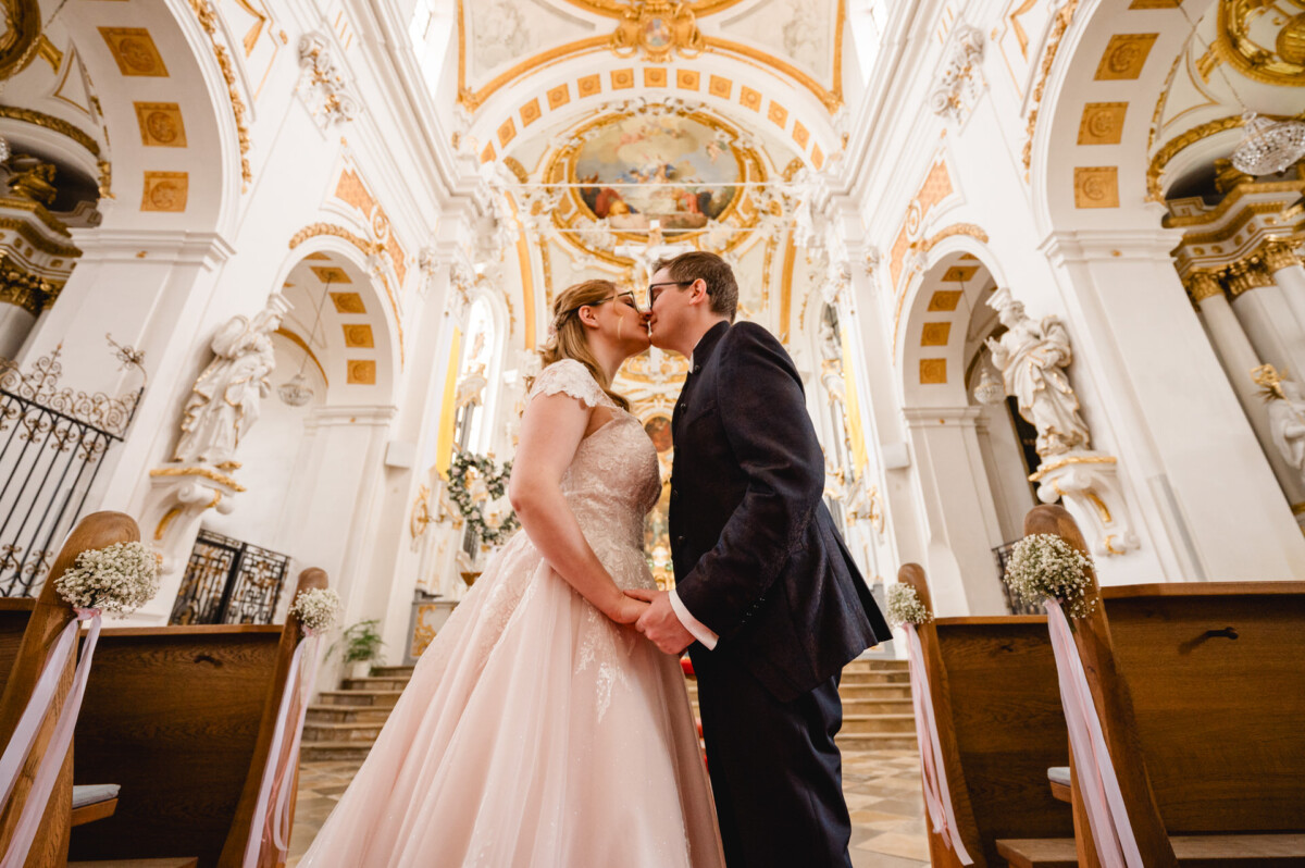 Ein Brautpaar küsst sich in einer geschmückten Kirche, während es zwischen Kirchenbänken und kunstvoller Architektur am Altar steht.