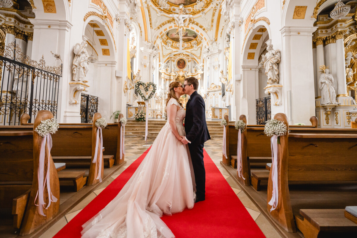 Ein Paar in Hochzeitskleidung steht auf einem roten Teppich in einem geschmückten Kirchengang, umgeben von kunstvoller Architektur und Holzbänken.