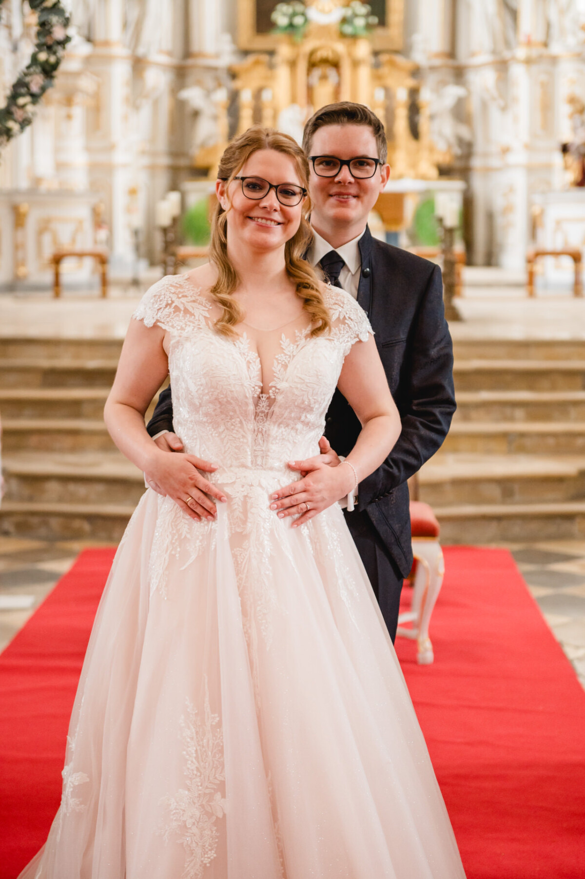 Eine Braut im weißen Kleid und ein Bräutigam im Anzug stehen lächelnd auf einem roten Teppich in einer dekorativen Kirchenumgebung.