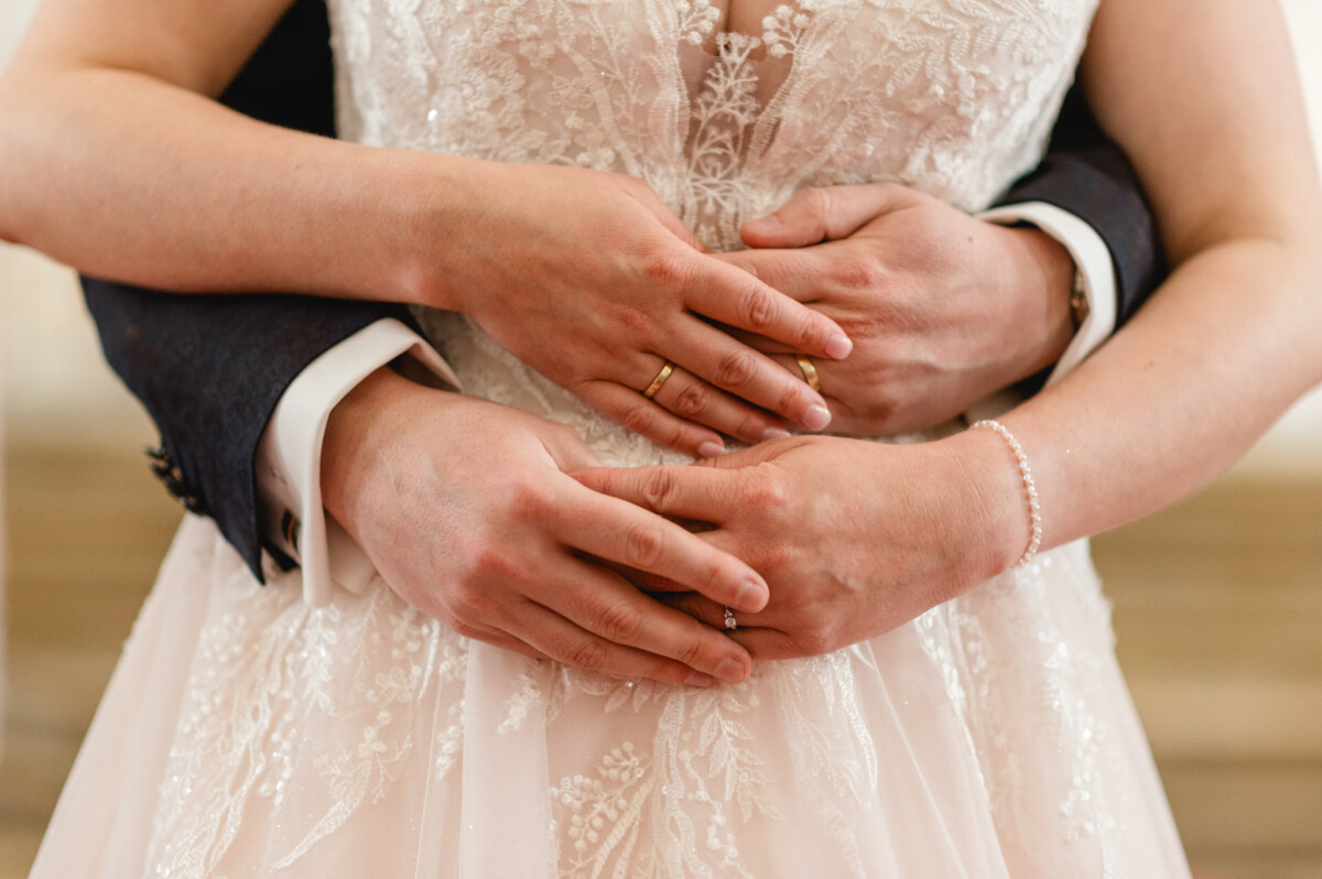 christian-lorenz.net Ein Paar in Abendgarderobe umarmt sich und hebt seine Hände mit Eheringen vor einem Spitzenbrautkleid hervor.