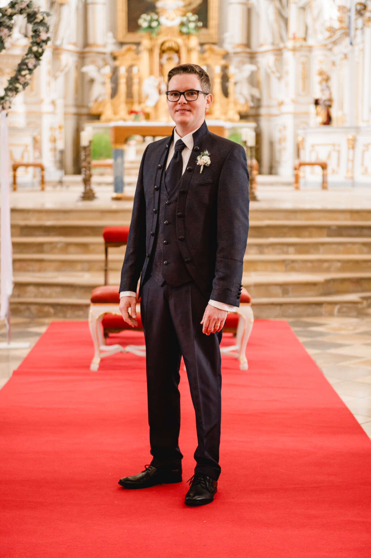Eine Person im Anzug steht auf einem roten Teppich in einer großen Halle mit kunstvollem Dekor und Stufen, die im Hintergrund zu einem Altar führen.