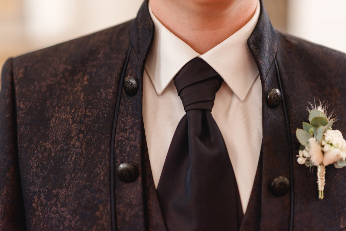 christian-lorenz.net Nahaufnahme einer Person, die ein dunkel gemustertes Jackett, eine schwarze Krawatte und ein weißes Hemd mit einer kleinen Ansteckblume am linken Revers trägt.