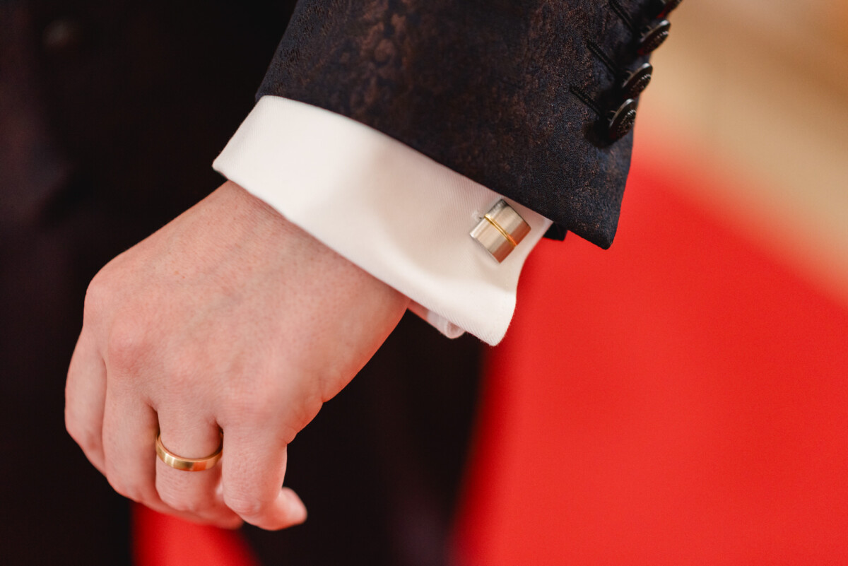 christian-lorenz.net Nahaufnahme der Hand einer Person, die einen goldenen Ring trägt. Die Hand ruht neben einem Anzugärmel mit einer weißen Manschette und einem silbernen Manschettenknopf vor einem roten Hintergrund.