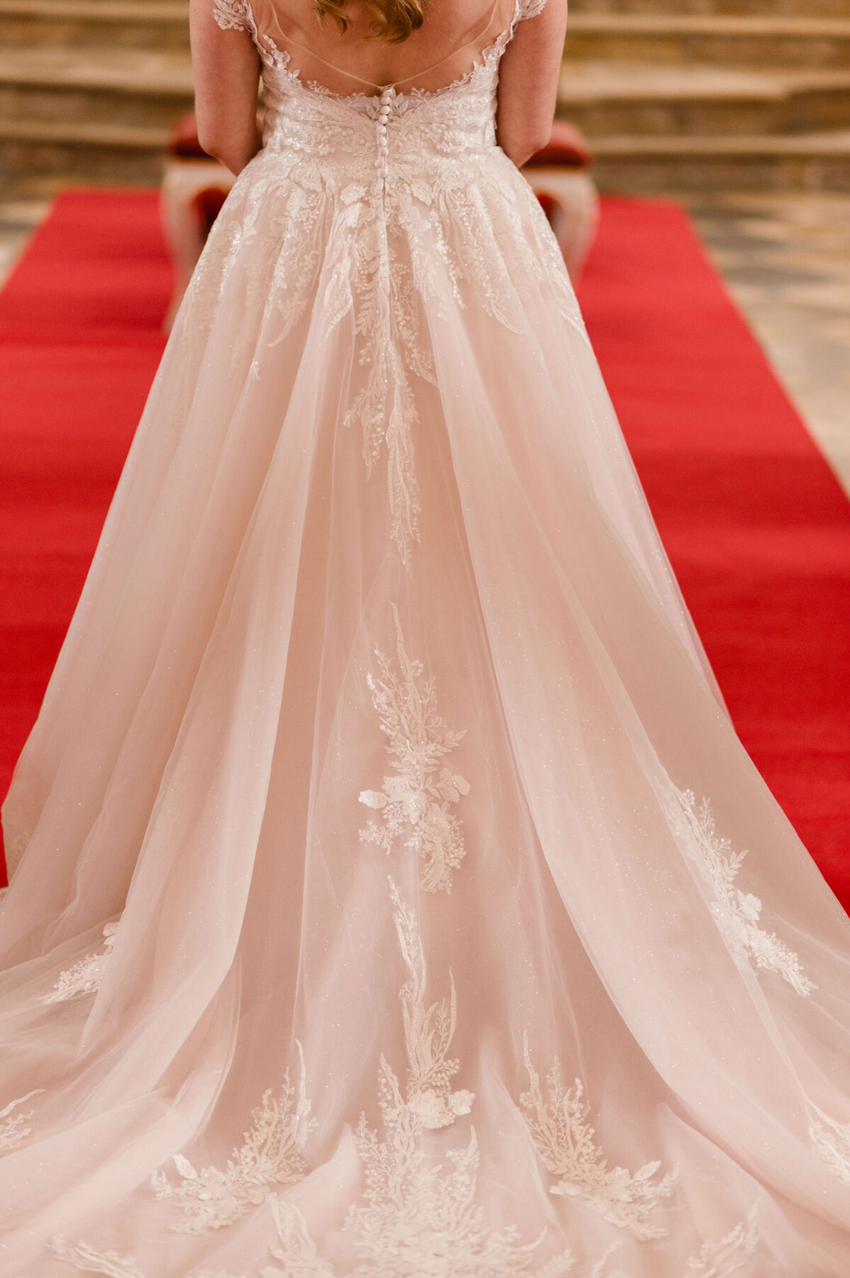 christian-lorenz.net Rückansicht einer Braut in einem kunstvollen weißen Kleid mit Spitzendetails und langer Schleppe, die auf einem roten Teppich steht.