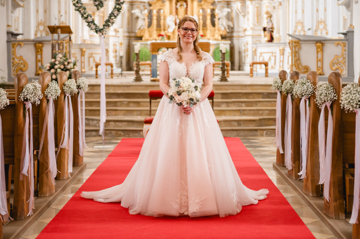 Eine Braut in einem weißen Kleid steht auf einem roten Teppich in einer geschmückten Kirche, hält einen Blumenstrauß und lächelt in die Kamera.