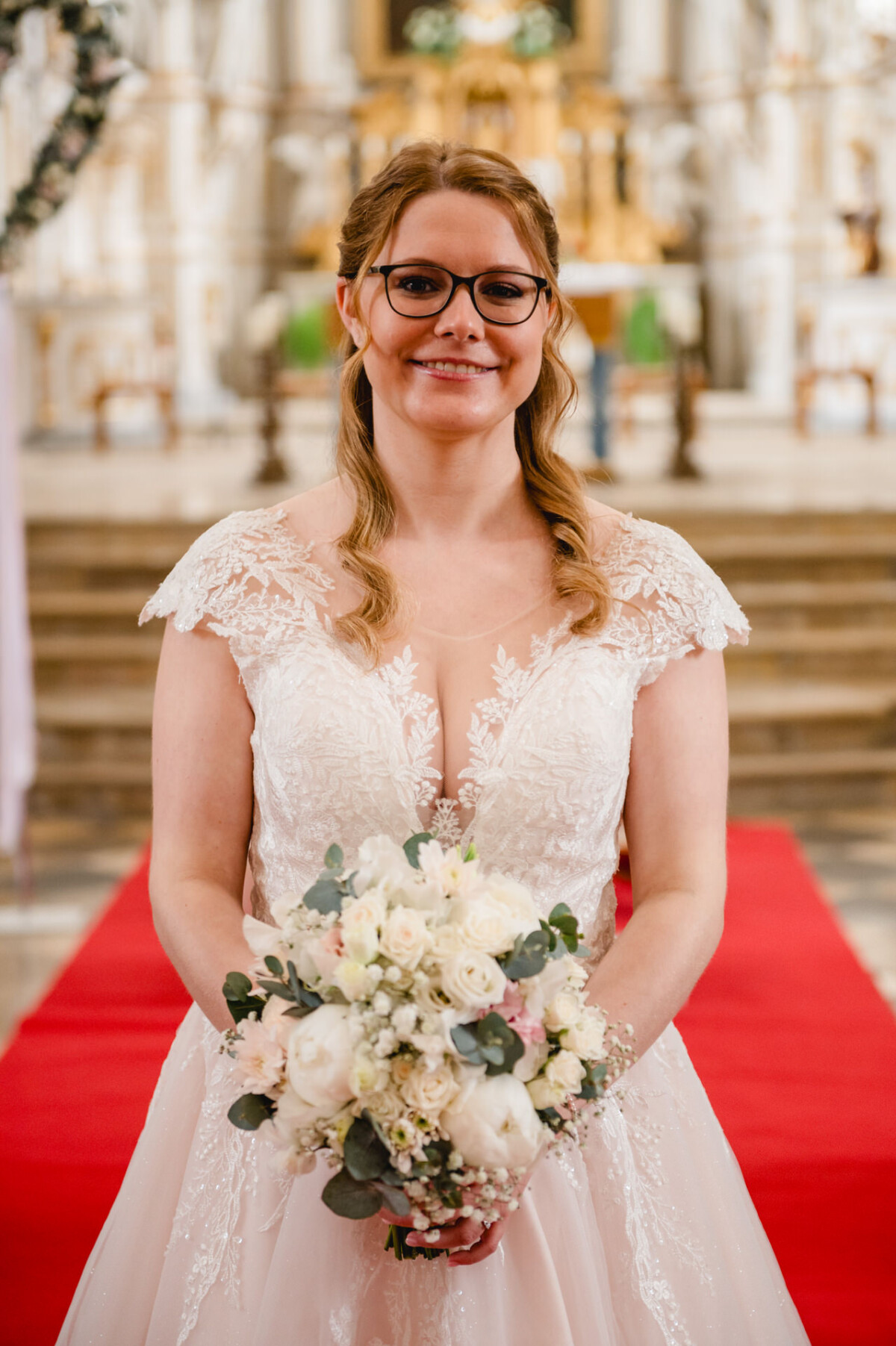 Braut in einem weißen Spitzenbrautkleid mit einem Strauß weißer Blumen in der Hand, steht auf einem roten Teppich in einer Kirche.