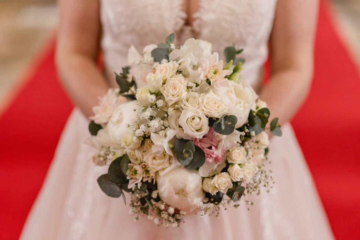 christian-lorenz.net Braut hält einen Strauß weißer und blassrosa Blumen mit Grün und steht auf einem roten Teppich.