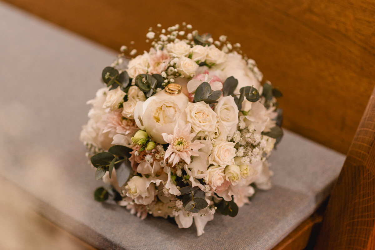 christian-lorenz.net Ein Brautstrauß mit weißen und rosa Rosen, Grünpflanzen und Schleierkraut, platziert auf einer grauen Bank.