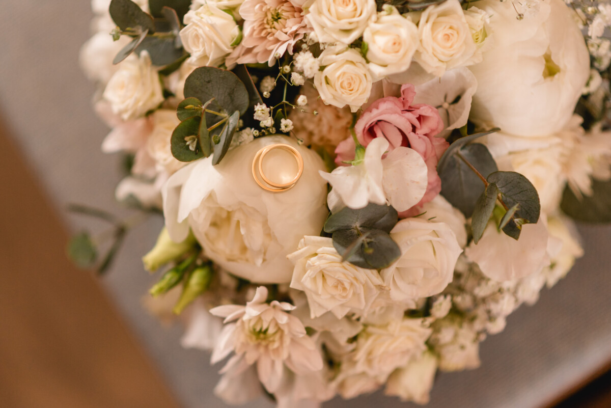 christian-lorenz.net Ein Strauß weißer und rosa Blumen mit zwei goldenen Ringen oben drauf, umgeben von Grün.